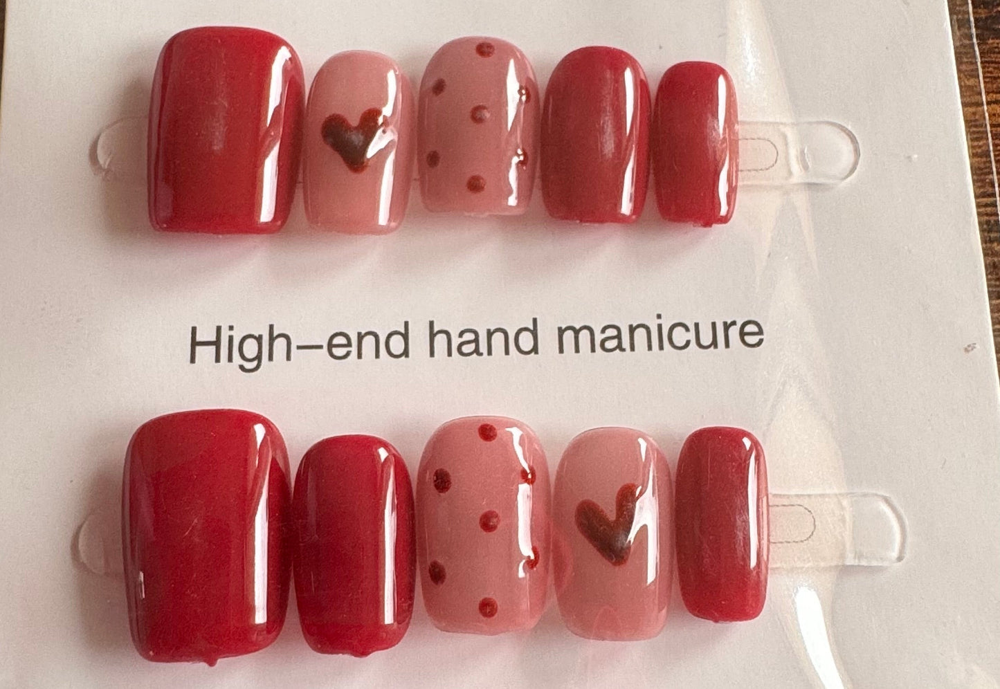 Set of red nail polish with heart designs on a labeled card, placed on a wooden surface.